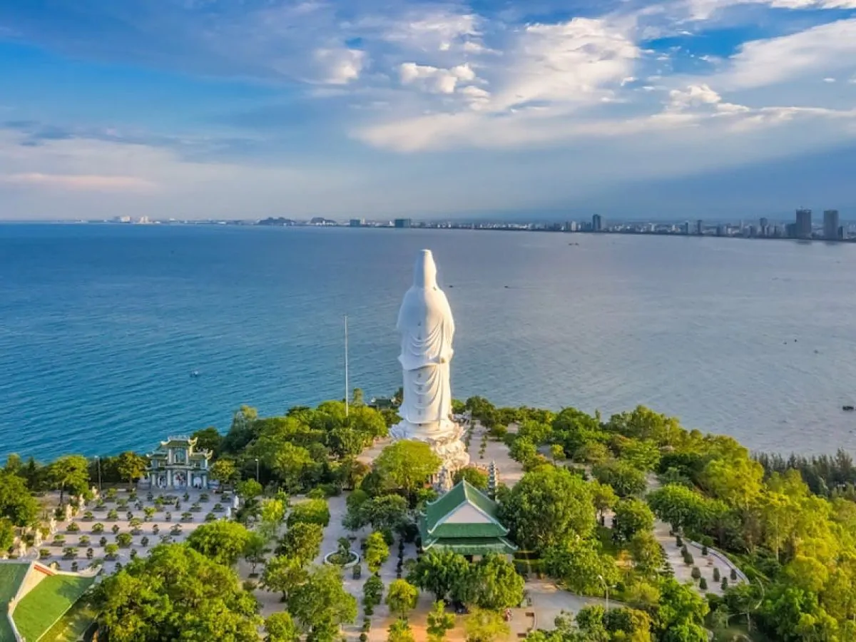 Aerial view of the 67-metre Lady Buddha statue at Linh Ung Pagoda on Son Tra Peninsula Da Nang, surrounded by tropical forest with Da Nang Bay and city skyline in the background