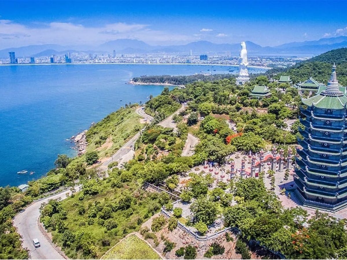 Aerial panorama of Son Tra Peninsula Da Nang showing the multi-tiered pagoda tower, winding coastal road with vehicle, Linh Ung temple complex and Da Nang city skyline with Marble Mountains in the background
