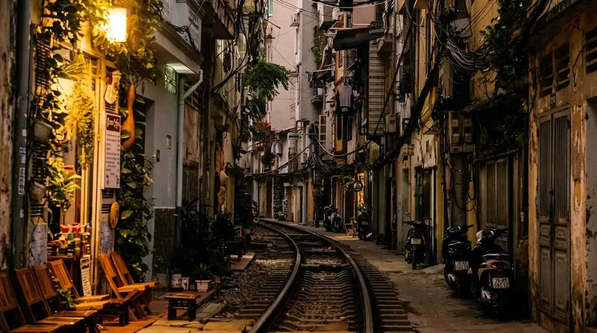 Quiet Hanoi Train Street alley at dusk with warm lantern lights, railway tracks, and old houses