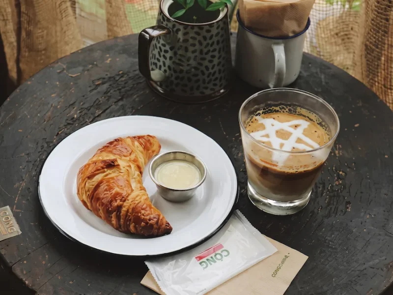 Vintage Interior of Cong Caphe Nha Tho Street - Vietnam Discovery Tours A cup of coffee decorated with a white star pattern served with a croissant and condensed milk at Cong Caphe Hanoi