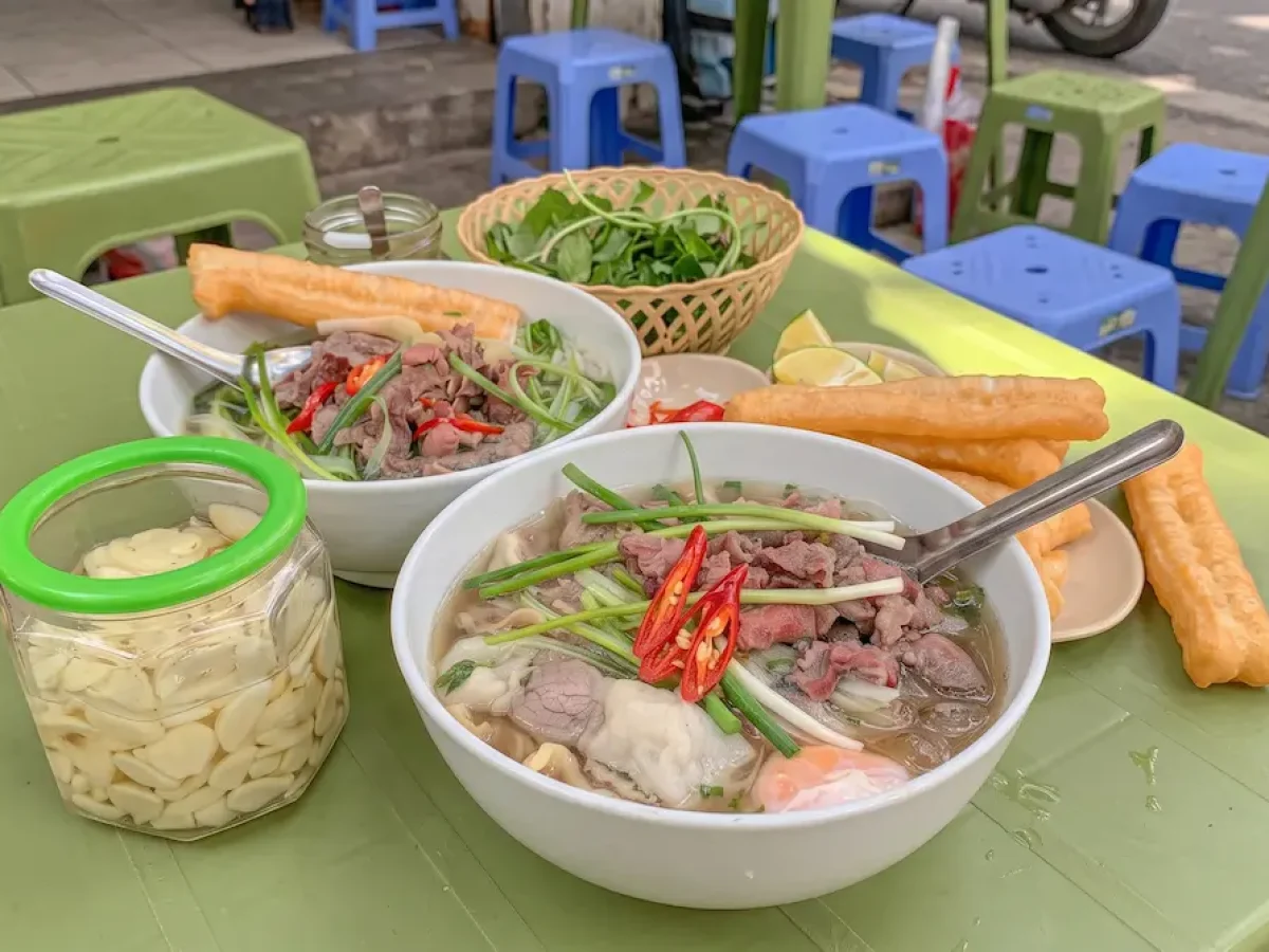Authentic Pho Hanoi Street Food - Vietnam Discovery Tours Two bowls of authentic Pho Hanoi with beef, fresh herbs, and fried dough sticks on a green street food table.