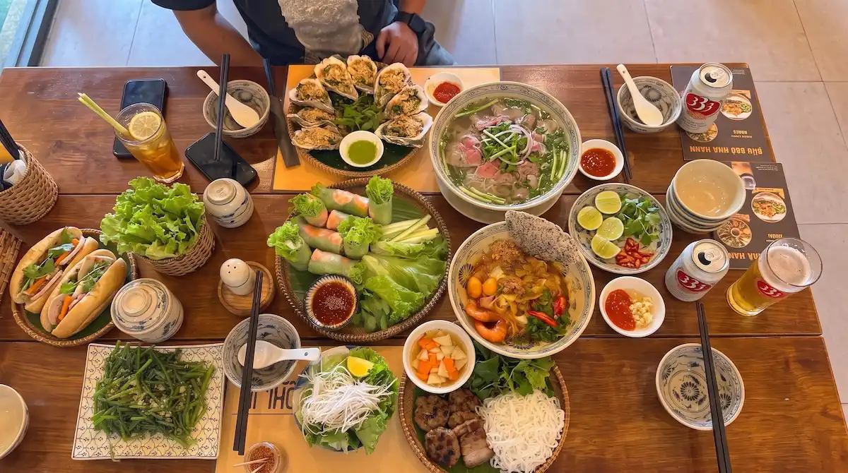 Authentic Vietnamese Food Feast - Vietnam Discovery Tours Top view of a wooden table filled with authentic Vietnamese dishes including Pho, Banh Mi, fresh spring rolls, Bun Cha, and local 333 beer.