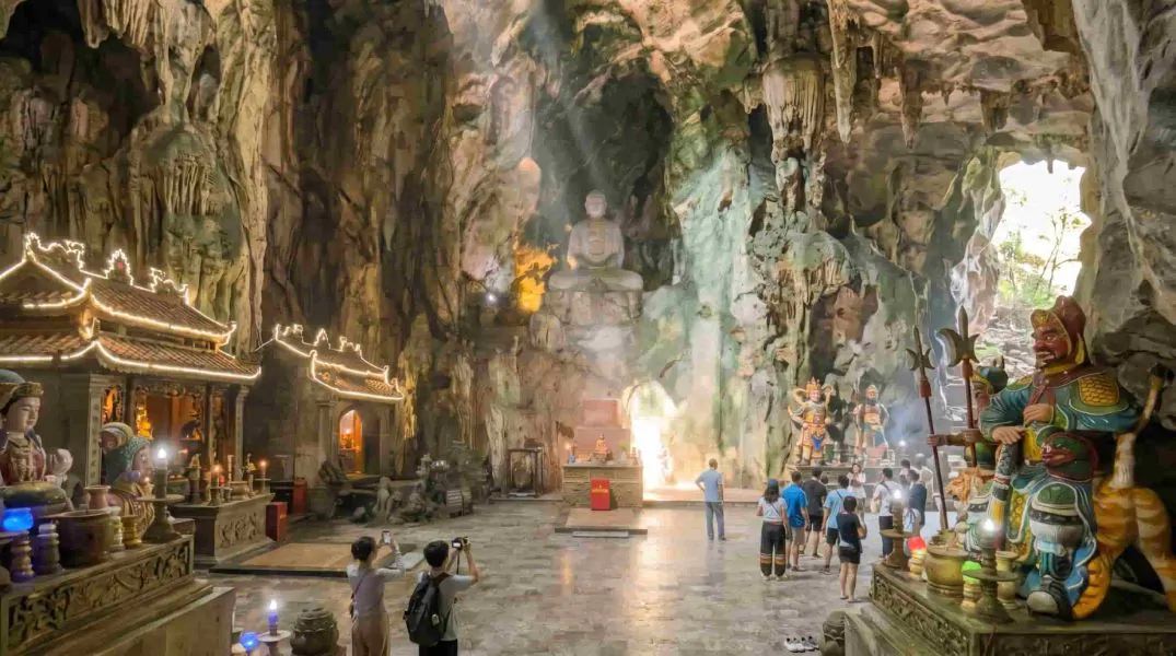 Interior of Huyen Khong Cave inside Marble Mountains Da Nang showing natural skylight beam, illuminated temple altar, giant Buddha statue and visitors exploring the sacred cave