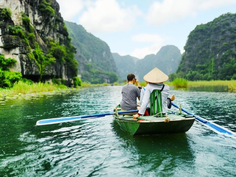 Trang An river boat ride A rower in a conical hat taking tourists down a river between tall limestone karst cliffs in Ninh Binh
